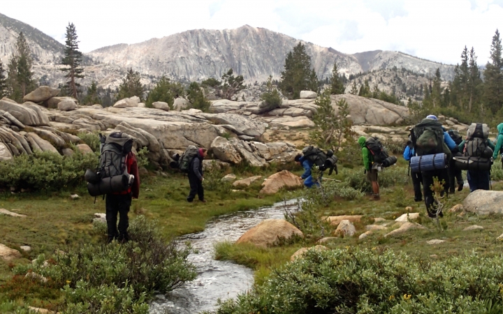 A group of outward bound students wearing backpacks hike beside a stream in a grassy field. There are mountains in the distance.